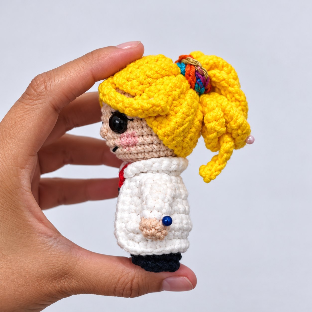 Detalhe de boneca de amigurumi com jaleco branco, cabelo loiro preso e acessório colorido